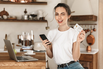 Young student girl sitting indoors using laptop computer chatting by mobile phone holding credit card.