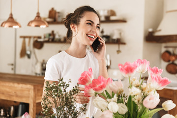 Beautiful happy young student girl posing indoors near flowers talking by mobile phone.