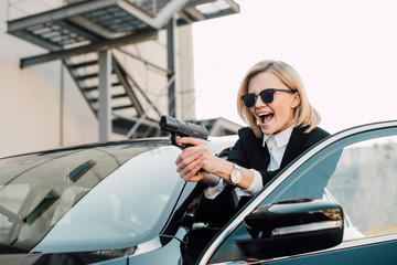 Fototapeta na wymiar cheerful blonde woman in sunglasses holding gun near black car