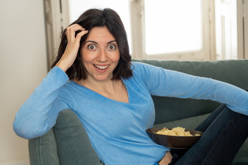 Lifestyle portrait of cheerful young woman sitting on the couch watching TV holding remote control