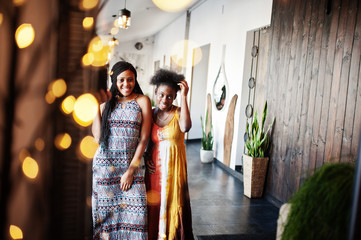 Two black african girlfriends at summer dresses posed in cafe at mirror.