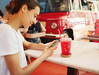 pretty young tanned woman in white t-shirt is sitting in cafe and uses phone, looking, reading or searching something. The glass with watermelon lemonade on the foreground and red vintage bus on