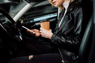 cropped view of girl holding paper cup and using smartphone in automobile