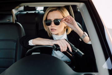 Obraz na płótnie Canvas beautiful young blonde woman holding gun at touching sunglasses in car
