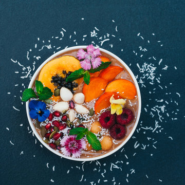 Isolated shot of bowl with tropical fruit decorated with flowers, mint, coconut flakes isolated over dark blue background. Fresh persimone, raspberry and almond.