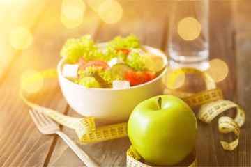 Salad in white bowl, fresh green apple and tape measure on dark wooden background. Diet and weight loss concept.