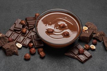 glass bowl of chocolate cream or melted chocolate, pieces of chocolate and hazelnuts on dark concrete background