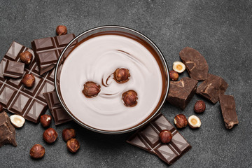 glass bowl of chocolate cream or melted chocolate, pieces of chocolate and hazelnuts on dark concrete background