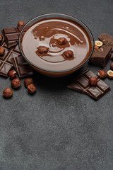 glass bowl of chocolate cream or melted chocolate, pieces of chocolate and hazelnuts on dark concrete background