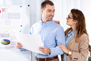 Obraz premium cheerful woman in glasses looking at handsome man with blank paper near white board with charts and graphs