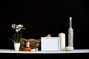 picture frame mockup. Shelf against black wall with decorative candle, glass, wood and rocks. Home plant orchid in pot.
