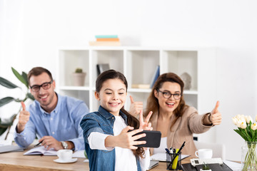 selective focus of cheerful child taking selfie on smartphone near happy parents gesturing in office