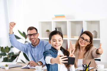 selective focus of cheerful kid taking selfie on smartphone near happy parents gesturing in office