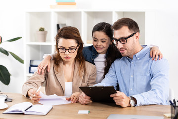 cheerful kid hugging mother and father in glasses looking at papers in office