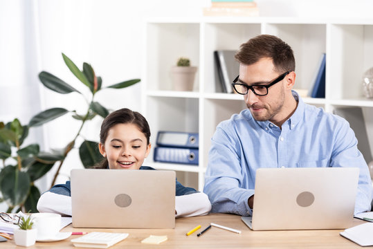 handsome father looking at laptop near cute daughter in office