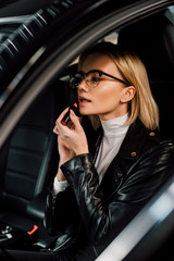 Fototapeta na wymiar beautiful blonde girl in glasses applying lipstick in car