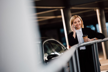 Fototapeta na wymiar selective focus of cheerful blonde girl talking on smartphone and holding paper cup near black car