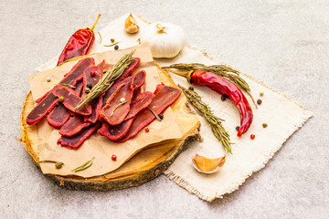 Basturma, dried tenderloin of beef meat, jerky, thinly sliced. Dry rosemary, pepper mix, chili, garlic on vintage linen cloth. Delicious food on a wooden board, stone background, close up.
