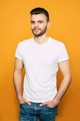 Stature and well-built man in denim jeans and white basic T-shirt with a beard and smooth dark hair is standing in front of orange background.