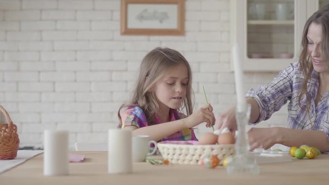 Cute little smiling girl painting Eastern eggs with a small brush sitting at the table with her mom in the kitchen. Preparation for Easter holiday. Mother and daughter have fun together at kitchen