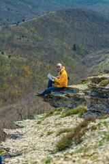 A tourist in a yellow jacket and glasses works on a laptop while on vacation in the mountains. In the background mountains. Freelance concept.