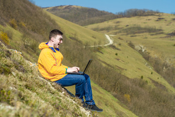 A tourist in a yellow jacket and glasses works on a laptop while on vacation in the mountains. In the background mountains. Freelance concept. Emotion of delight