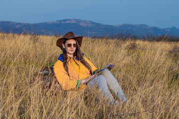 A young girl with long dark hair in a yellow jacket, leather cowboy hat and glasses sits in the same grass, leaning on a tourist backpack, looking into the distance. Background mountains, sky.