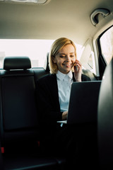 Fototapeta na wymiar smiling blonde woman talking on smartphone and looking at laptop in car