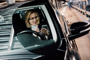 emotional girl in glasses showing middle finger while looking at smartphone in car