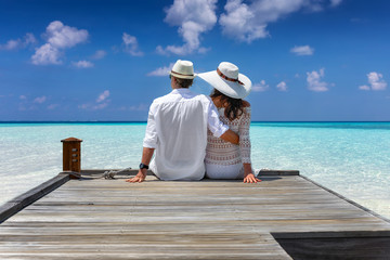Elegantes Paar in weißer Sommerkleidung in den Flitterwochen auf den Malediven sitzt auf einem Holzsteg und genießt die Aussicht 