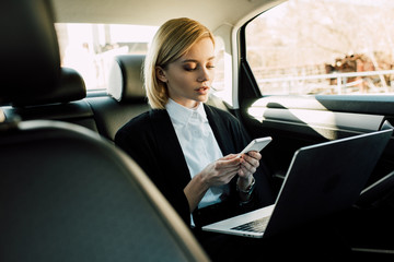 attractive blonde young woman looking at smartphone while sitting with laptop in automobile