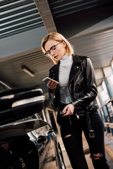 serious and attractive girl holding smartphone and key near black car