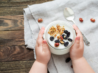 Natural homemade yogurt in a glass jar. Healthy food for breakfast in child's hands. Kid holding jar with muesli on linen tablecloth on on wooden table.