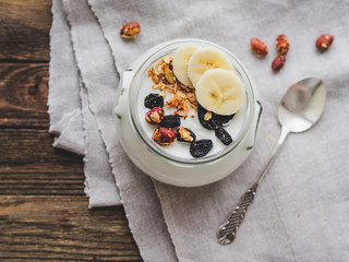 Natural homemade yogurt in a glass jar. Healthy food for breakfast with muesli. Jar with granola and banana slices on linen tablecloth on on wooden table.