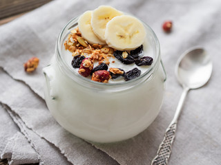Natural homemade yogurt in a glass jar. Healthy food for breakfast with muesli. Jar with granola and banana slices on linen tablecloth on on wooden table.
