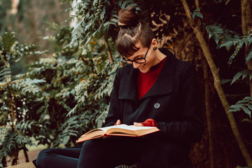 Young attractive elegant woman in eyeglasses reading volume and sitting on bench in city garden