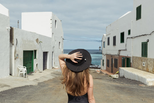 Back view of stylish woman touching hat while standing on shabby street of small coastal town on cloudy day near sea