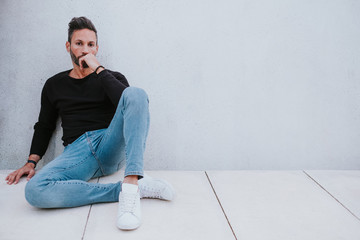 Adult handsome elegant pensive male in casual wear sitting on floor and looking at camera near grey wall