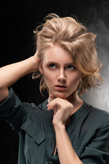 Beautiful young blonde girl with disheveled hairstyle and nude makeup, wearing a shirt and jeans emotionally posing on gray background. She is looking thoughtfully up and holding her head.