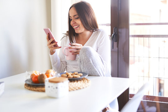 Attractive young happy woman using mobile phone and having breakfast at table near window at home