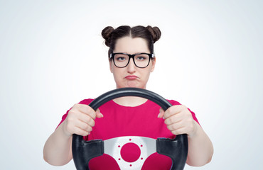 Unhappy young girl in glasses with steering wheel, on light background. Front view. Female car driver concept