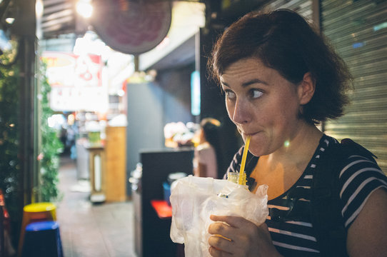 Funny female traveler sucking fresh beverage from bottle and looking away while standing near cafe on city street in Thailand