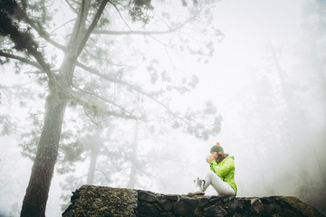 Mountain loving woman drinking cold coffee, sitting on a stone wall resting after trekking in the woods immersed in the mist. Concept of wellness and outdoor life. Warm tones autumn natural landscape