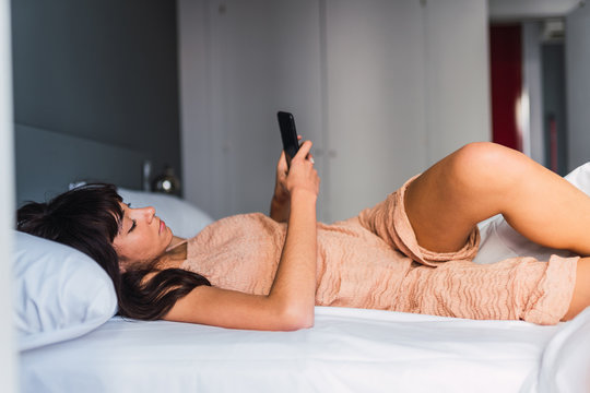 Side view of young woman using mobile phone and resting on bed