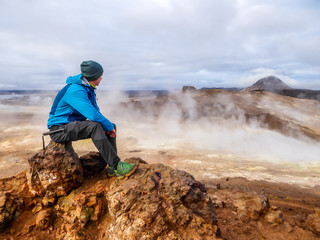 Young man wearing a blue jacket sits on top of a mud mountain, overlooking a geothermal spot noted for its bubbling pools of mud & steaming fumaroles emitting sulfuric gas. Power of planet Earth.