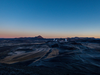 A volcanic, geothermal landscape captured during the sunrise. First beams of the sun reach the top parts of volcano. Smoke coming out of hot springs. Volcanic lava fields all around. Cosmic and alien