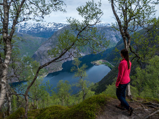 Obraz premium A girl wearing pink shirt stands at the edge of the hill and supports herself on a tree, while looking down to the fjord valley. Stunning view. Fjord going deep into the valley. Hiking remedy.
