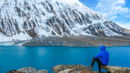 Naklejka premium Young man sitting at the rock in front of Tilicho Lake. Blue and calm surface of the lake, mountains covered in the shadow, sunlight in the back. Overcast. Harsh landscape. Thankfulness, freedom.