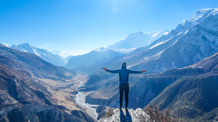 Man wearing a beanie and blue jumper, spreads his arms wide, breathing deeply the fresh mountain air. His gesture represents freedom and happiness. Below a long valley stretches in Himalayas. - obrazy, fototapety, plakaty