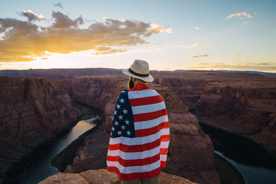 Back view of male wrapped with USA flag standing near beautiful canyon against sunset sky on West Coast